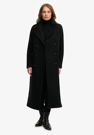 Lange zwarte wollen jas met dubbelgeknoopte voorkant, grote revers en twee zijzakken. Draagt over een zwarte coltrui en broek.