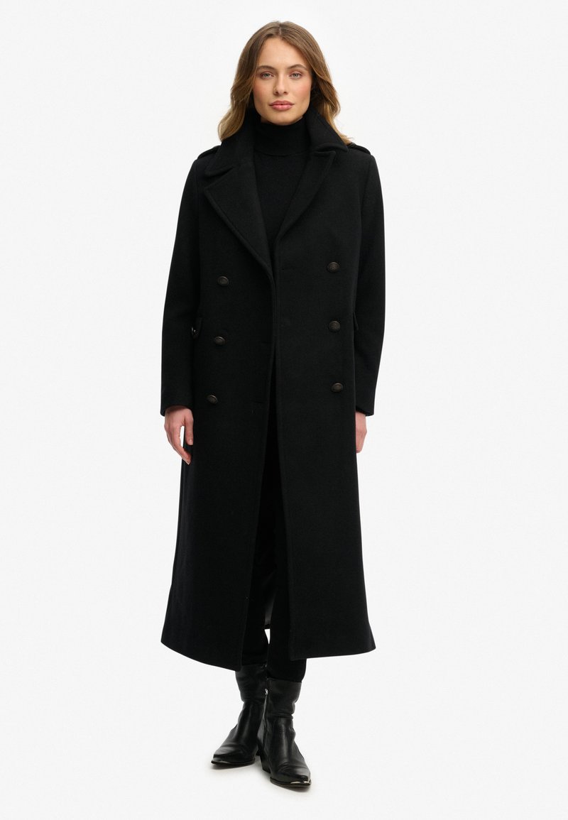 Lange zwarte wollen jas met dubbelgeknoopte voorkant, grote revers en twee zijzakken. Draagt over een zwarte coltrui en broek.