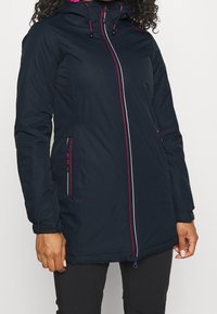 Femme portant une veste à capuche bleu marine avec des détails de fermeture éclair rose et blanc et des poches zippées, associée à un pantalon noir.