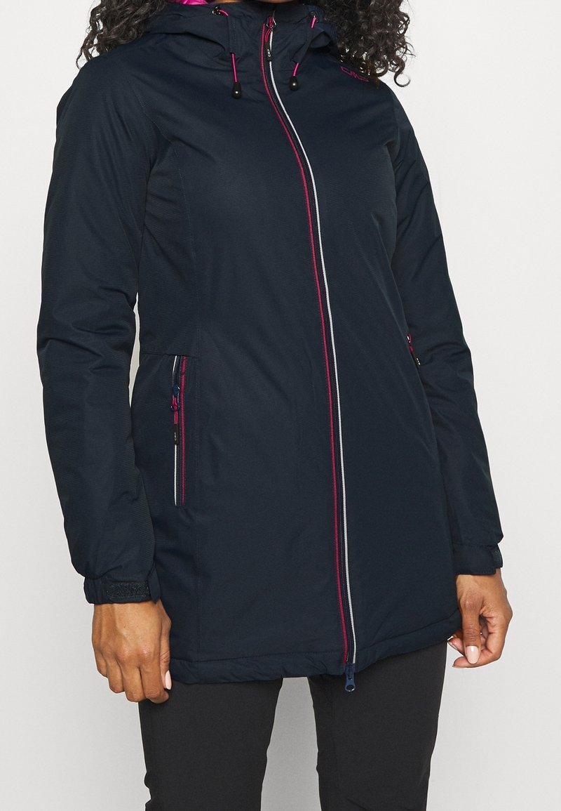 Femme portant une veste à capuche bleu marine avec des détails de fermeture éclair rose et blanc et des poches zippées, associée à un pantalon noir.