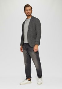 Szary blezer w wzór houndstooth na jasnoszarym swetrze, w połączeniu z ciemnoszarymi dżinsami i białymi sneakersami. Prezentuje nowoczesny, casualowy look.