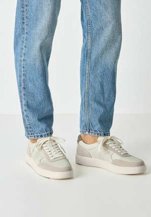 Baskets gris clair et beige avec des textures en cuir lisse et en daim, présentant des accents perforés et des lacets plats, associées à un jean en denim bleu.