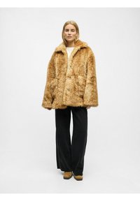 Nepluizijn in warm goudbruin, oversized pasvorm met een brede kraag, sluiting met knopen aan de voorkant en grote zijzakken. Gecombineerd met zwarte broek.