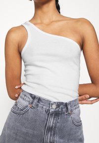 Débardeur blanc côtelé avec une découpe sur une épaule, affichant une coupe ajustée. Associé à un jean en denim gris délavé taille haute.