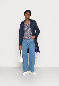 Casaco de trench navy sobre uma blusa floral, jeans azul claro e ténis brancos. A segurar uma mala branca acolchoada de forma arredondada.