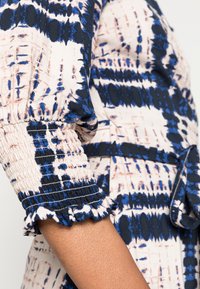 Manche avec poignet élastiqué et détail à volants sur tissu à motifs aux tons bleus, noirs et beiges, portée sur un bras.