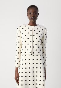 White textured jacket and skirt set with large black polka dots. Jacket features a rounded neckline and front closure.