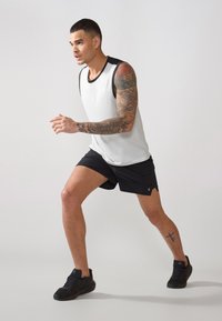Weißes ärmelloses Sporttop mit schwarzen Akzenten, kombiniert mit schwarzen Shorts. Das Modell trägt schwarze Sportschuhe und zeigt Tätowierungen an Armen und Beinen.