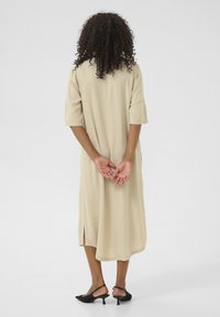 Robe beige longueur genoux avec des manches courtes, fentes latérales et texture lisse. Portée avec des talons slingback noirs. Vue de l'arrière.