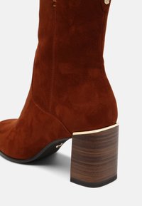 Bottine marron en daim avec un bout arrondi, une forme effilée et un talon bloc présentant une texture grain de bois et un accent métallique.