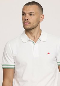 Polo shirt blanche en coton, dotée d'un col et de poignets à rayures vertes. Un petit logo en forme de diamant rouge est placé sur la poitrine.