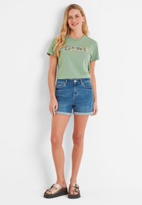 Green cotton t-shirt with colorful text, paired with blue denim shorts featuring rolled hems. Model wears brown slide sandals.