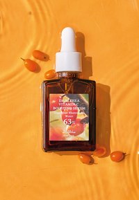 Botella gotero de vidrio marrón con tapa blanca. La etiqueta presenta un diseño colorido, dice "Suero Booster de Vitamina C", incluye porcentaje e ingredientes. Fondo naranja.