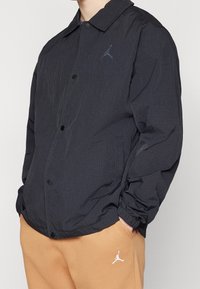 Chaqueta ligera negra con una textura arrugada, cierre de botón y un pequeño logo de Jordan en el pecho; combinada con pantalones color beige que presentan un logo.