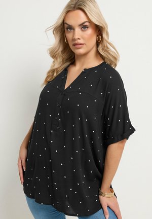 Blonde woman wearing gold hoop earrings and bracelets, dressed in a loose black shirt with white polka dots and blue jeans, posing calmly.