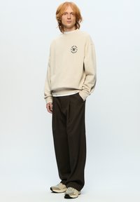 Beige sweatshirt met rond logo, losse donkerbruine broek en beige sneakers. Model staat tegen een effen lichte achtergrond.
