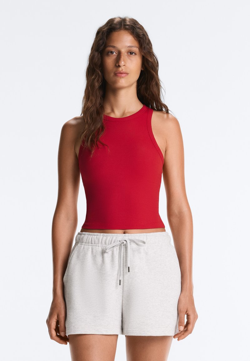 Woman with long wavy hair wearing a sleeveless red crop top and light gray drawstring shorts standing against a plain background.