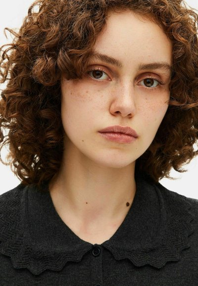 Maglione di lana nera con colletto a merletto e chiusura con bottoni. Il modello ha capelli ricci marroni e lentiggini, con uno sfondo neutro.
