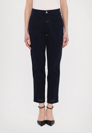 Vrouw draagt nauwsluitende donkerblauwe broek en zwarte enkellaarsjes met spitse neus en enkelriempje, staand tegen een effen witte achtergrond.