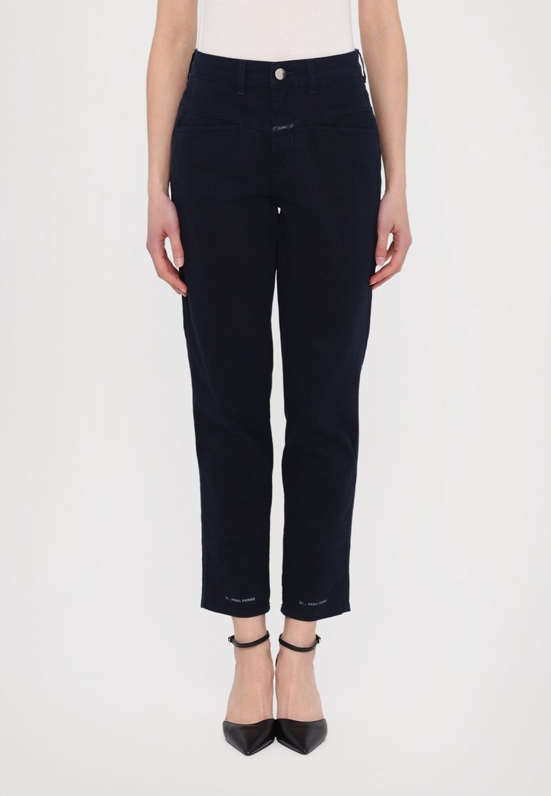 Femme portant un pantalon slim bleu marine foncé et des talons noirs à bride à la cheville et bout pointu, debout devant un fond blanc uni.