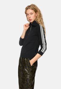 Schwarze Zip-up-Strickjacke mit gerippter Textur, die weiße Streifen an den Ärmeln hat. Kombiniert mit glänzenden, grünen Leopard-Print-Hosen.