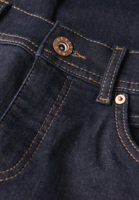 Jeans de mezclilla azul oscuro con herrajes de bronce, costuras doradas en contraste y una superficie texturizada. Presenta un cierre de botón de metal y trabillas para cinturón.