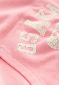 Tissu rose avec une texture lisse présentant de grandes lettres blanches fissurées qui indiquent "DIS". Coutures visibles et zones de matériel doux et extensible.