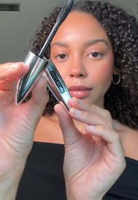 Tube de mascara argenté au design effilé, étiqueté "FAUX CILS", tenu par une main, avec une brosse en poils naturels. Surface lisse et réfléchissante.