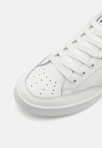 Fehér sneaker sima bőr felsőrésszel, kerek orral, perforált részletekkel és lapos fehér fűzőkkel. A talpa textúrázott gumiból készült.
