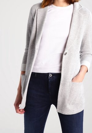 Femme portant un blazer ouvert gris clair par-dessus un t-shirt blanc et un jean bleu foncé, avec une main dans la poche du blazer.