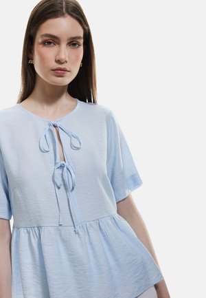 Blouse bleu clair à manches courtes, avec un col rond et un détail à nouer à l'avant. Tissu texturé, présentant une forme peplum ample.