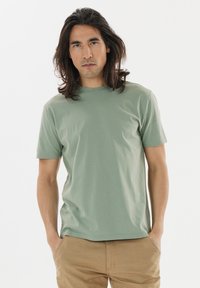 Short-sleeve, crew-neck green t-shirt made of soft cotton. Model stands with hands in pockets, wearing light brown pants.