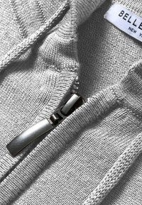 Primo piano di un tessuto grigio a maglia con una cerniera argento e un'etichetta di abbigliamento parzialmente visibile sul colletto.