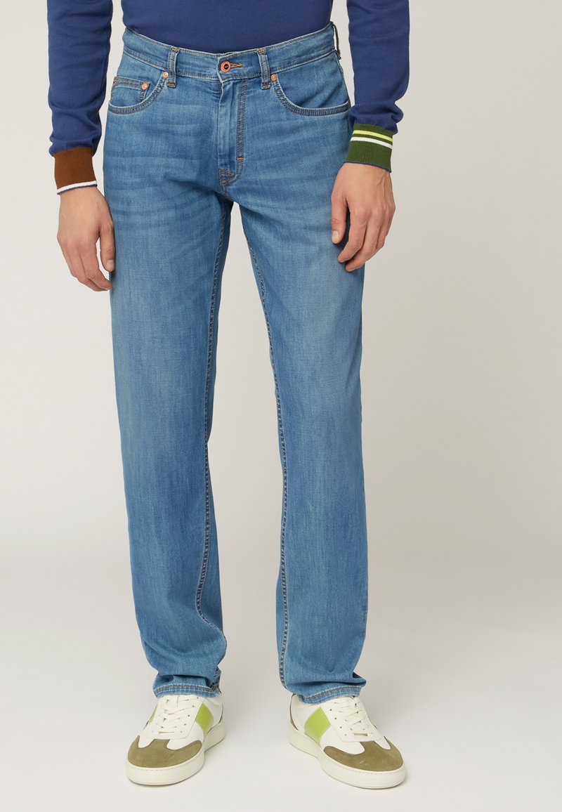 Jeans in denim blu chiaro con vestibilità rilassata, caratterizzati da dettagli cuciti, passanti per cintura e tasche frontali, abbinati a sneakers bianche e verdi.