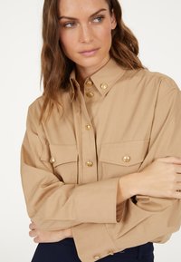 Tan button-up shirt with gold hardware, featuring two chest pockets and a structured collar, made of lightweight fabric with a smooth texture.