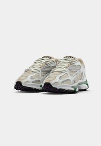 Une paire de sneakers Lacoste blanches et beiges avec des panneaux en mesh, des semelles épaisses, des accents verts et violets, et des lacets blancs sur un fond uni.