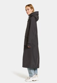 Lång mörkgrå regnjacka med huva, med dragkedja framtill, fickor och elastiska ärmslut. Bärs över avslappnade ljusblå jeans och sneakers.