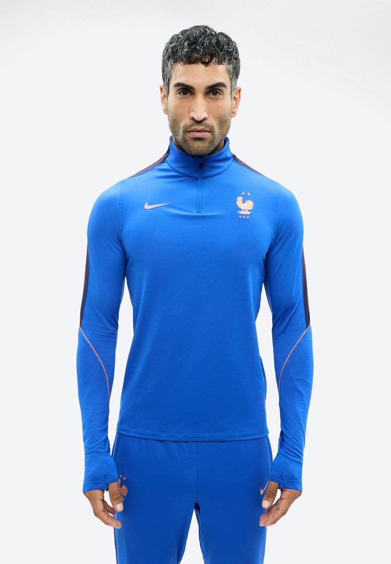 Mees, kes kannab sinist Nike spordiriietust Prantsuse Jalgpalliliidu logoga, seisab valge taustaga ja on näoga edasi.