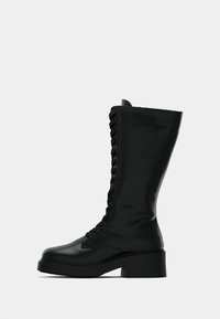 Μαύρη μπότα μέχρι το μισό της γάμπας με κορδόνια, με παχιά χαμηλή φτέρνα και λείο δερμάτινο υλικό, εμφανίζεται από το πλάι σε άσπρο φόντο.