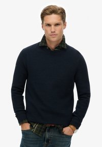 Navyblauer, strukturierter Strickpullover, der über einem grün-schwarz plaidierten Hemd getragen wird, mit rundem Halsausschnitt und gerippten Bündchen, kombiniert mit verwaschenen Jeans.
