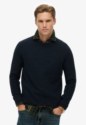 Suéter de punto texturizado en color navy, superpuesto a una camisa de cuadros verde y negro, con cuello redondo y puños acanalados, combinado con jeans desgastados.