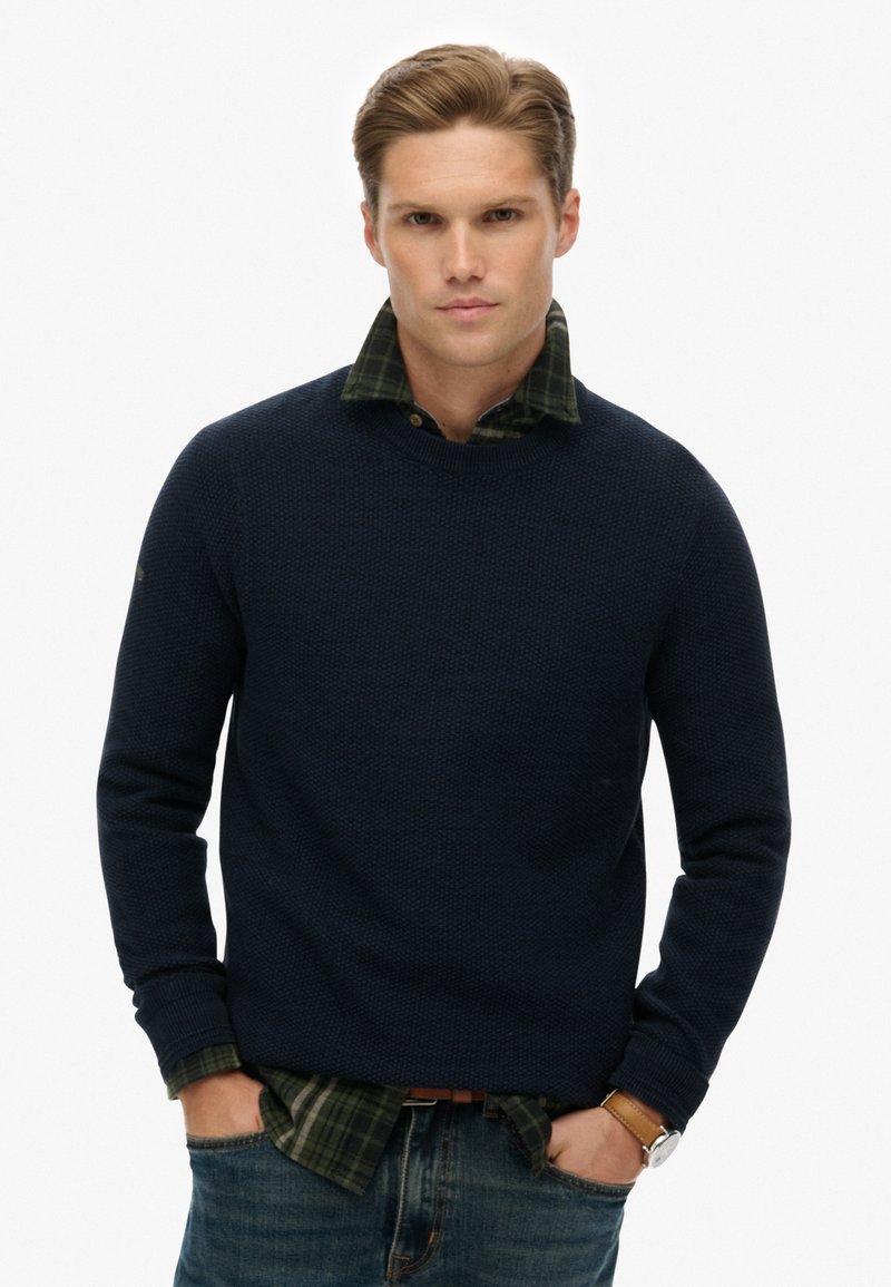 Navyblauer, strukturierter Strickpullover, der über einem grün-schwarz plaidierten Hemd getragen wird, mit rundem Halsausschnitt und gerippten Bündchen, kombiniert mit verwaschenen Jeans.