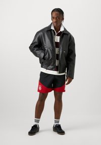 Μαύρο δερμάτινο μπουφάν με μπροστινό φερμουάρ και δύο πλάγιες τσέπες, φορεμένο πάνω από ένα άσπρο και μπορντό ριγέ πουκάμισο και μαύρο-κόκκινο αθλητικό σορτς.