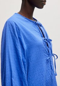 Blaue Baumwollbluse mit einem strukturierten, gewebten Design, runder Halslinie, Bindeschluss und langen Ärmeln mit lockerer Passform.
