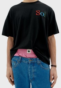 Schwarzes Baumwoll-T-Shirt mit mehrfarbiger "Sol"-Stickerei, kombiniert mit hellblauen Jeans. Sichtbarer pinker Bund mit Logo-Tag.