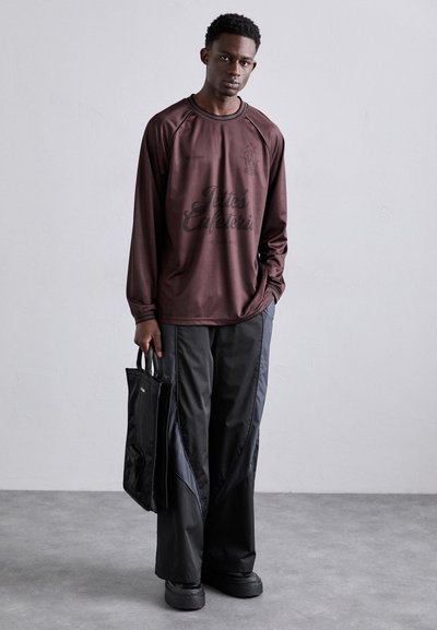 Long-sleeve brown shirt with graphic print, paired with black, loose-fitting, textured pants. Model holds a small black tote bag.