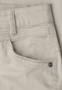 Pantalon beige clair avec poche avant, passant de ceinture visible, coutures doubles et détail de rivet en métal près du bord de la poche.