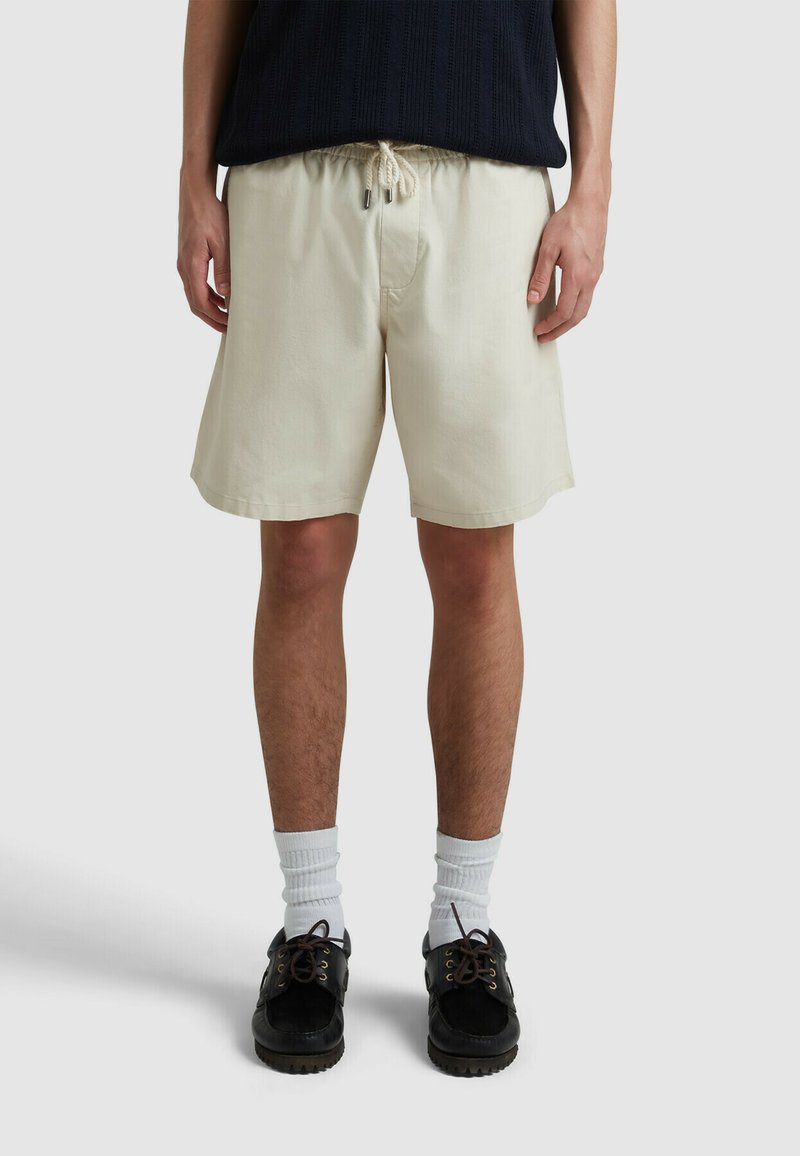 Shorts di cotone beige con vita regolabile, tasche laterali e vestibilità rilassata, abbinati a scarpe nere e calzini bianchi alla caviglia.