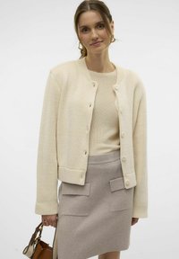 Cremefarbener Strickcardigan mit Rundhalsausschnitt und Knopfverschluss, getragen über einem beigen, gerippten Oberteil und einem hellgrauen, taillierten Rock mit Seitentaschen.