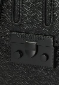 Bolso de mano de cuero negro texturizado con una placa de metal rectangular que lleva la inscripción "SEIDENFELT MANUFAKTUR," adornada con dos tachuelas hexagonales.